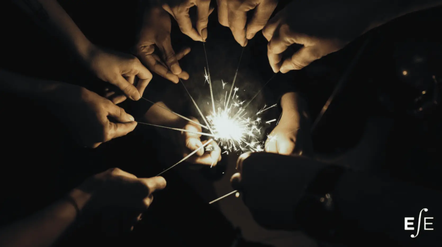 Close up of several sets of hands holding sparkler-style fireworks while another set of hands lights all of them at once
